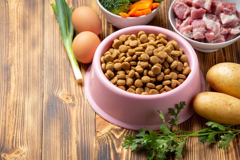 Dry dog food in a pink bowl surrounded by fresh raw ingredients including eggs, meat, vegetables, and potatoes on a wooden surface.
